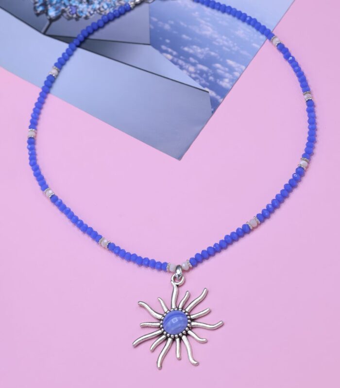 Photo of the "Azure Sun" necklace, which has white and purplish blue beads and a shiny sun charm, on a pink background.